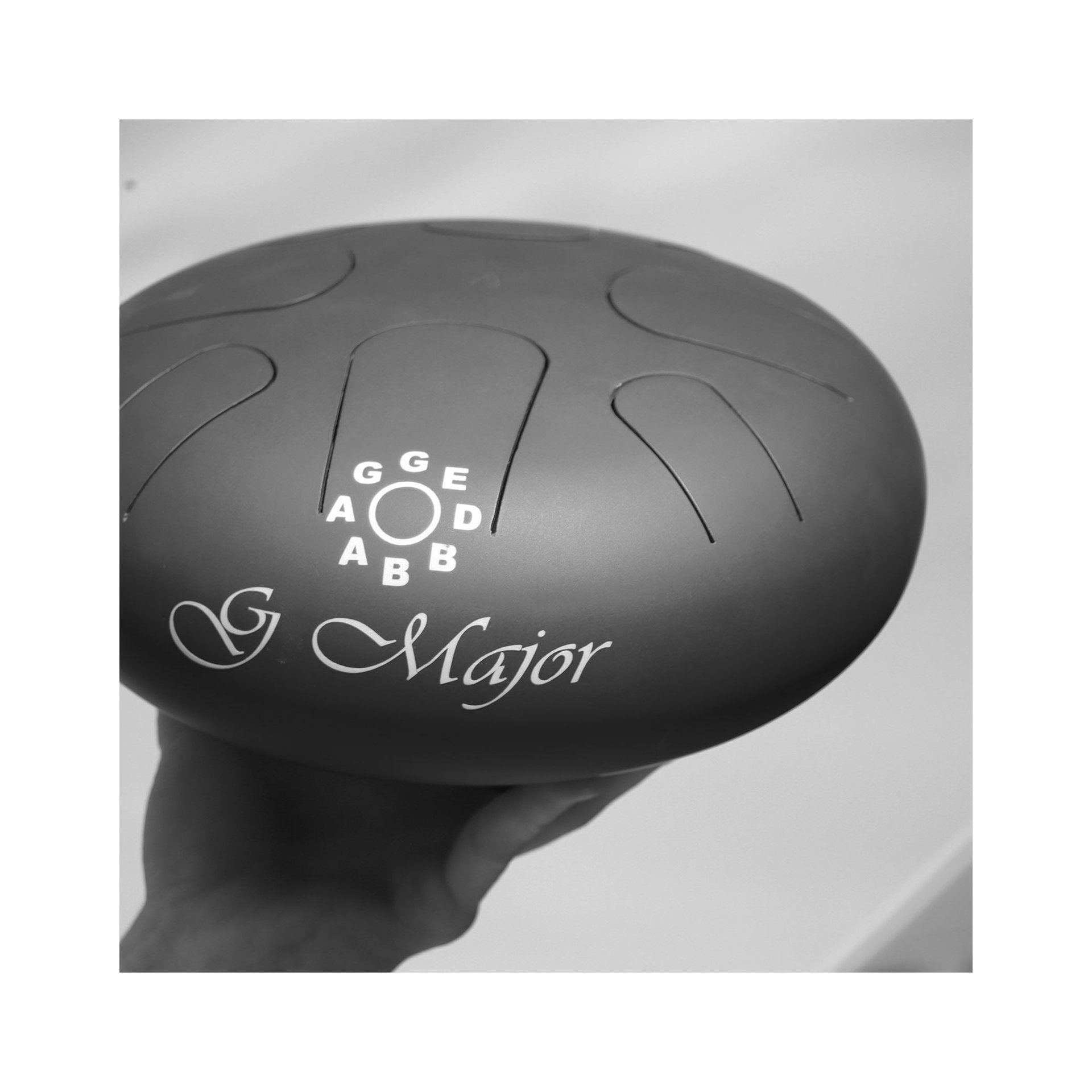 Side view of black steel tongue drum labeled 'G Major' being held in a hand, with note labels on top